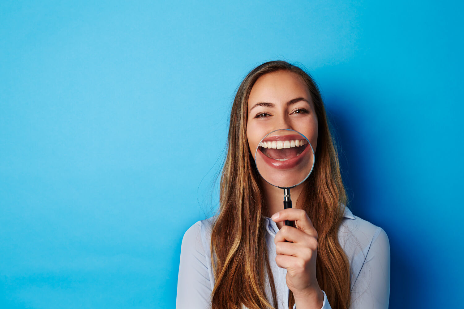 Brunette woman smiles and holds a magnifying glass to her teeth against a blue wall